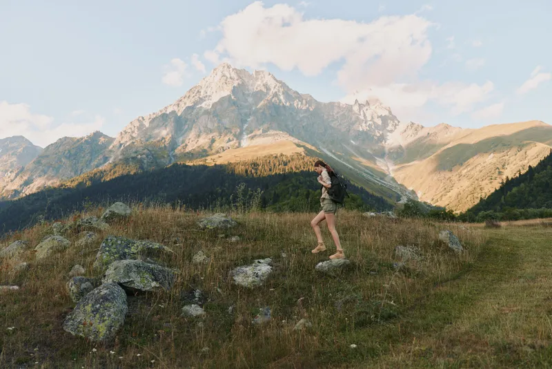A solo woman hikes along a grassy mountain ridge under a clear sky, with rocky slopes and distant peaks in view, wearing shorts and a light top, enjoying fresh air and freedom