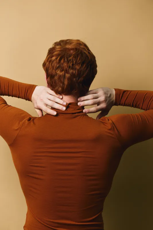 Person with red hair and pale skin wearing a brown long sleeve shirt seen from behind touching neck against beige background. Casual posture, minimalistic style, back view.