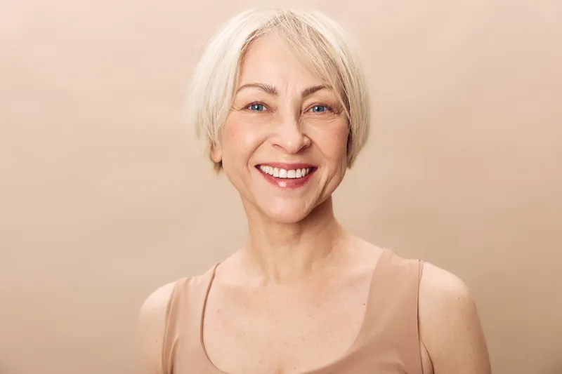 Smiling mature woman with short blonde hair wearing beige sleeveless top poses against plain background. Natural beauty, confidence, and positive mood concept for aging gracefully.