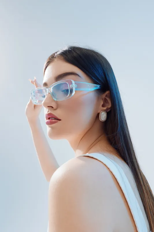 Young woman with long dark hair wearing transparent glasses and white top poses against a light background. Concept of beauty fashion style and modern accessories.