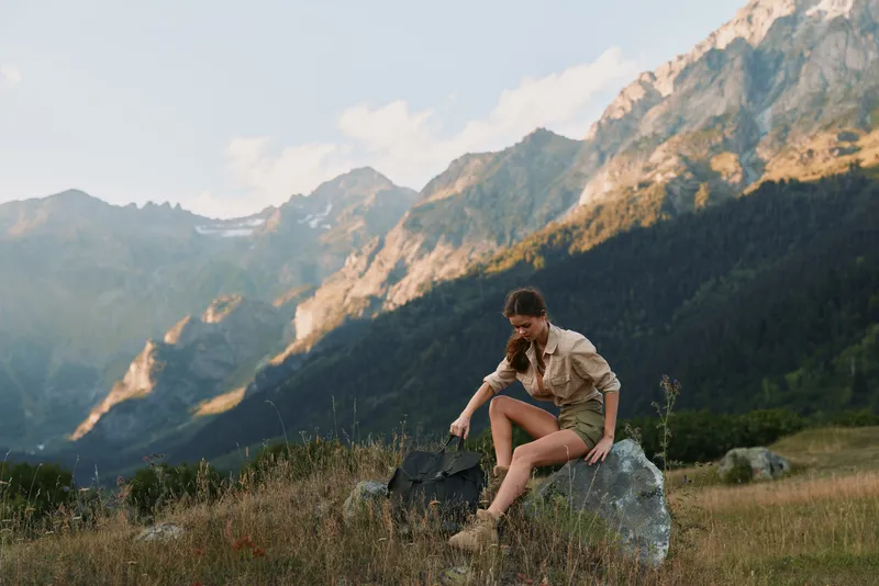 A young woman in casual clothing sits on a rock in a mountain meadow, adjusting her hiking boots during a sunny day. Concept of outdoor lifestyle and adventure.