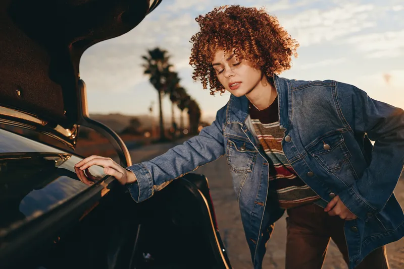 Woman lifestyle outdoors at sunset, curly hair, wearing denim jacket, leaning on car door, calm confident moment, hands on handle, warm light, soft sky, road background, city air