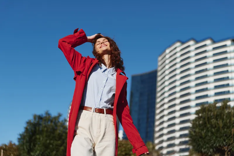 Young woman in a red coat and white shirt stands outdoors, looking up at the clear blue sky with a confident smile.