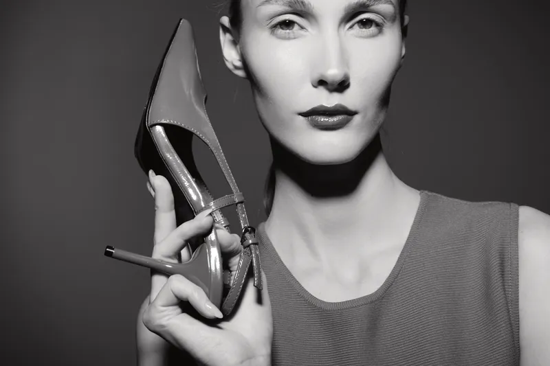 Portrait of a white woman with pale skin holding a high heel shoe near her face. Monochrome fashion image with elegant mood and focus on footwear and female beauty.