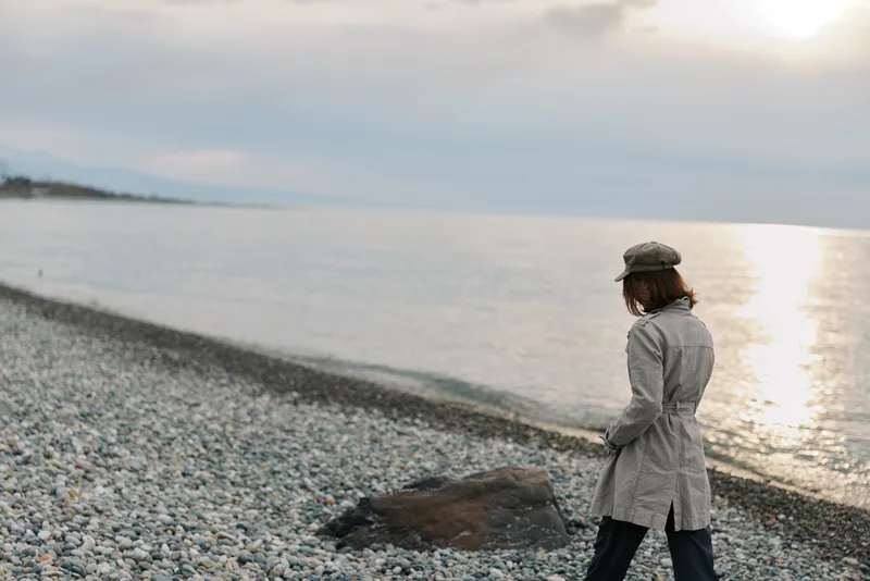 A woman in a grey coat and hat walks alone on a rocky beach near calm water under a cloudy sky. The muted colors and solitude create a contemplative mood and peaceful atmosphere.