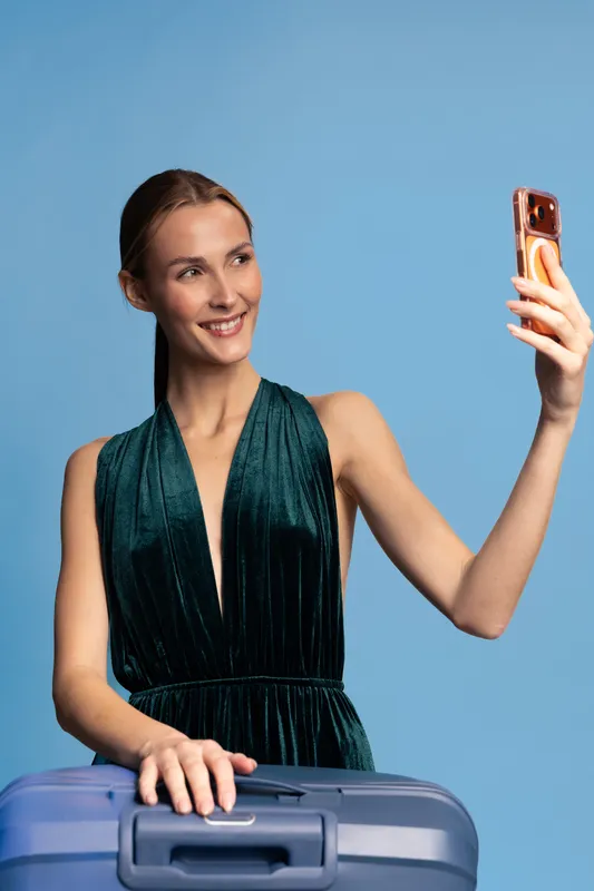 Smiling woman in green dress takes selfie with phone while holding blue suitcase on blue background. Concept of travel, vacation, tourism, and modern lifestyle with happy mood and leisure.