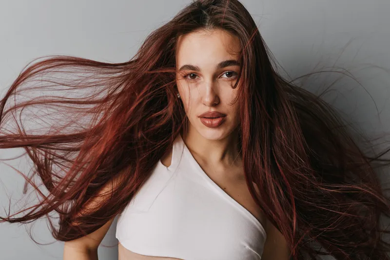 Woman with long flowing hair and natural makeup posing in white top against gray background. Portrait of confident female model with loose hair movement and direct gaze at camera.
