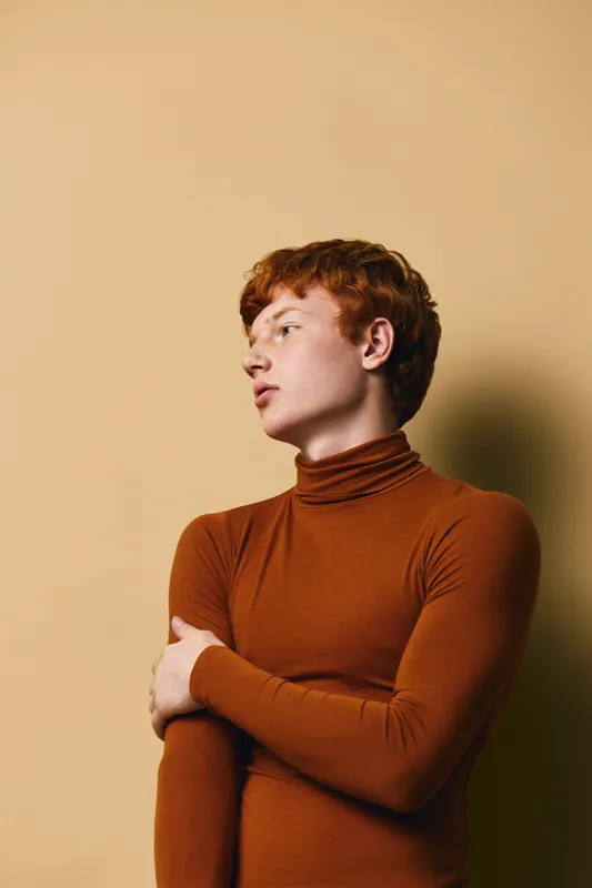 Young man with red hair wearing brown turtleneck sweater posing with arms crossed looking away against beige background. Portrait, fashion, style and casual clothing concept.