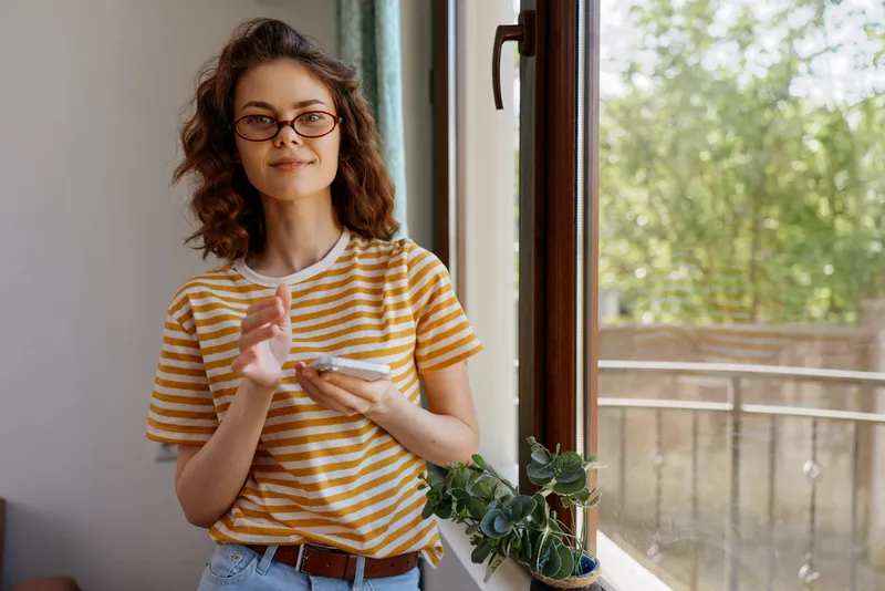 Young woman with curly hair and glasses stands by a window, holding a plate and spoon. Striped yellow shirt, casual home setting, natural daylight. Lifestyle moment featuring comfort, routine, and