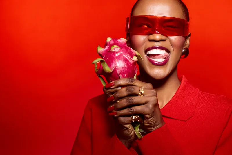 Smiling dark skinned African woman in red sunglasses and red outfit holds a dragon fruit against red background. Concept for healthy lifestyle, fruit, fashion, and vibrant colors.