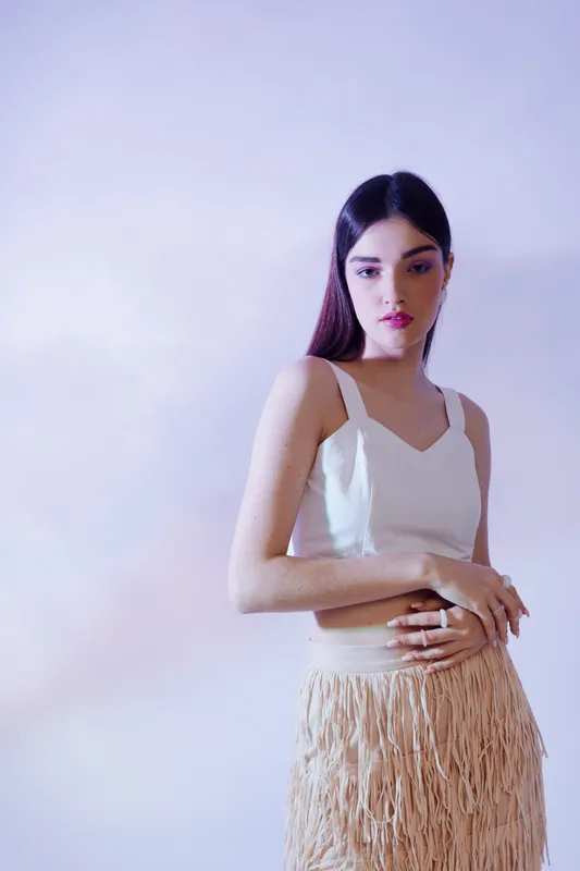 Young woman in a white top and beige skirt poses against a light background. The image highlights beauty, fashion, and elegance in a minimalist style.