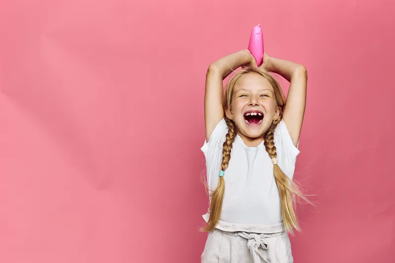 Happy girl with blonde braids holds a pink hairdryer above her head smiling widely on a pink background. Studio shot of playful childhood fun.