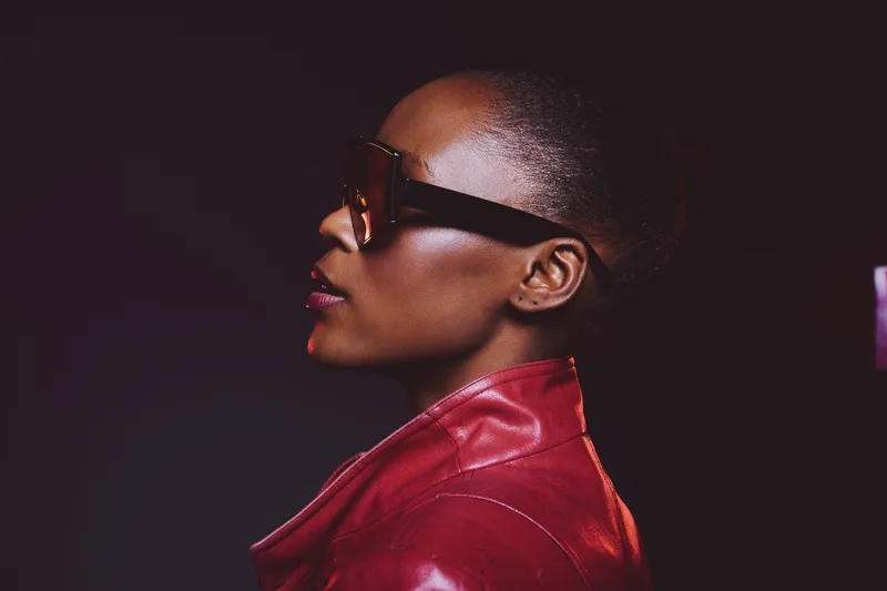 Profile of a confident black woman with dark skin wearing oversized sunglasses and a red leather jacket against a dark background. Concept of fashion, style, and urban elegance.