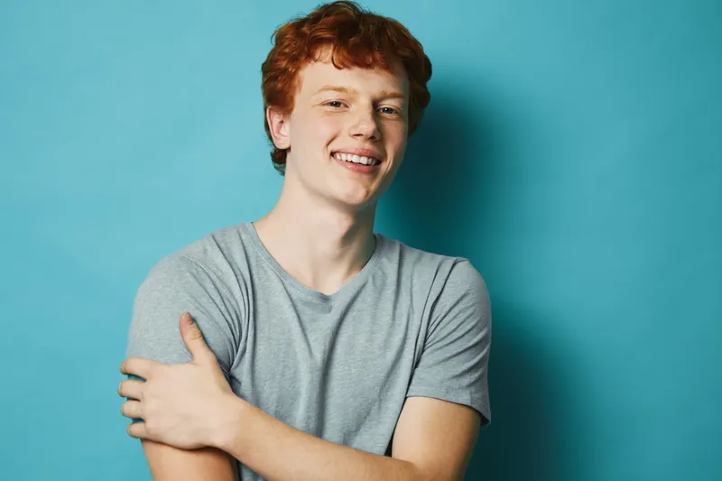 Portrait of smiling young man with red hair in casual grey t shirt standing with arms crossed in front of blue background, showing confident and relaxed expression.