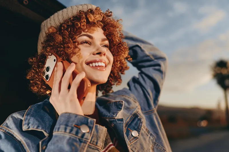 Smiling woman with curly hair talking on smartphone outdoors in a denim jacket, golden hour sunlight and soft wind, lifestyle moment with relaxed casual mood, near street today