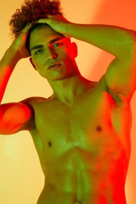 Young man with curly hair posing shirtless with hands on head under colorful red and green lighting for artistic portrait and fashion photography.
