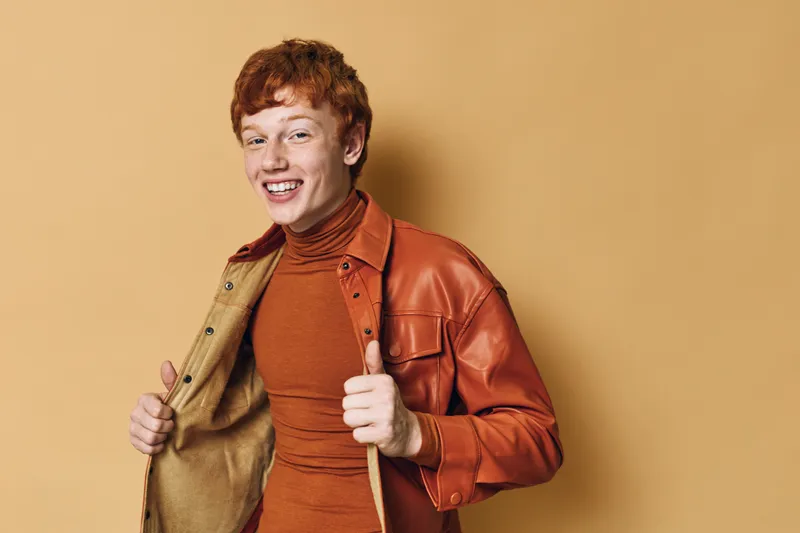 Young man with red hair wearing orange turtleneck and brown leather jacket smiling and posing on beige background. Casual style, confident, happy expression and modern outfit for fashion portrait.