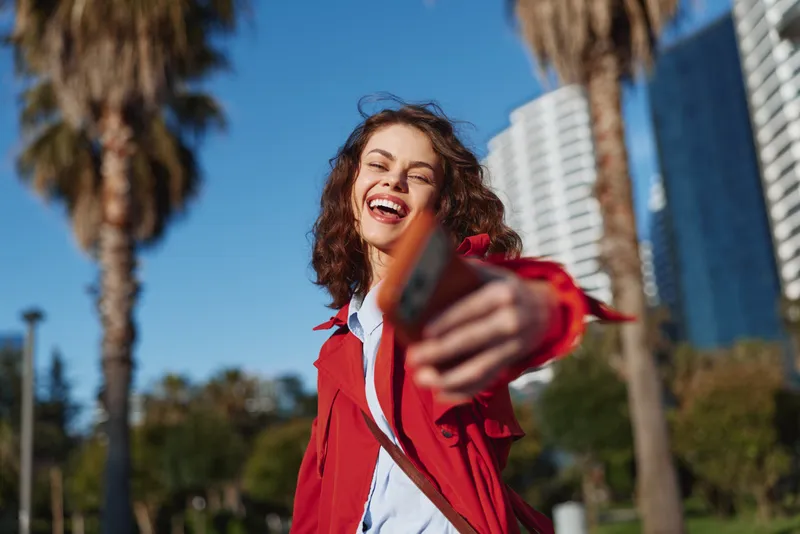 Young woman with curly hair poses outdoors, smiling as she takes a selfie in a sunny urban park with palm trees and tall buildings in the background under a bright blue sky.