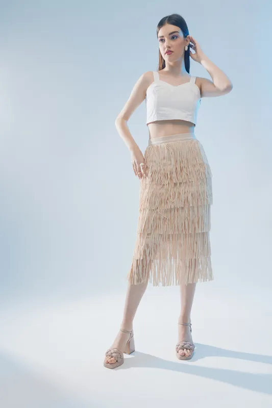 Young woman in a white crop top and beige fringe skirt poses confidently on a light background. Concept of beauty, fashion, style, and elegance in studio lighting.