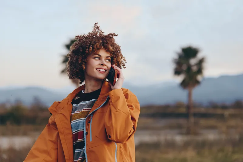 Woman lifestyle outdoors portrait in rural field, wearing orange jacket, curly hair, holding phone to her ear, smiling while talking, warm sunlight, sunset sky, blurred trees