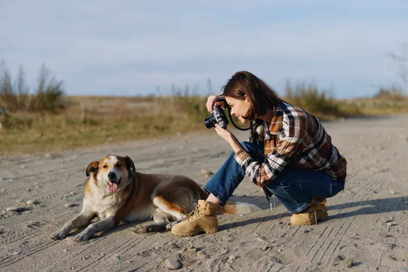Photographer kneeling beside a dog on a dirt road captures a candid outdoor moment with a handheld camera, sunny day, casual plaid shirt and denim boots.