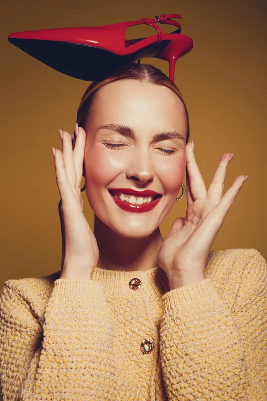 Smiling woman with closed eyes wears red high heel shoe on head, dressed in yellow knitted sweater. Studio portrait with warm lighting, concept of fashion, fun, and playful mood.