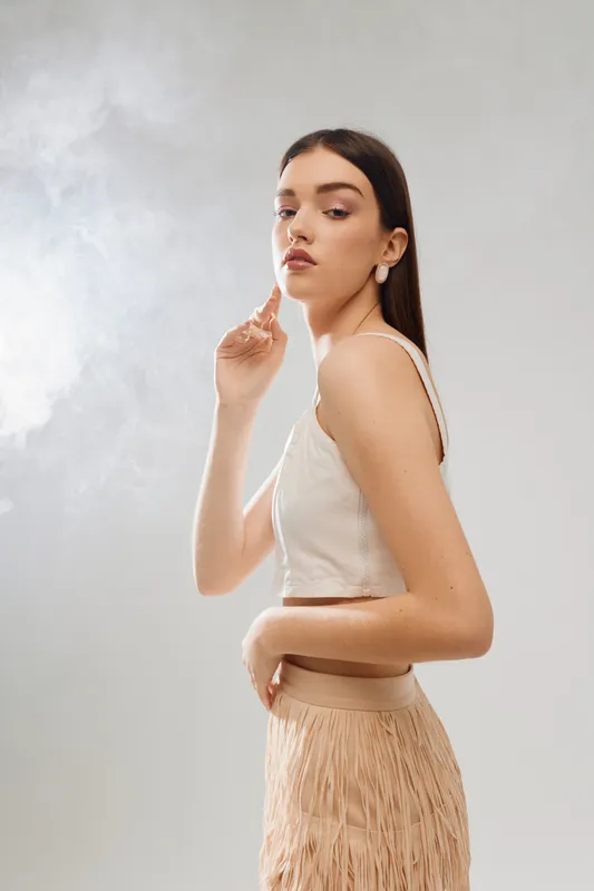 Young woman beauty fashion model posing in a white sleeveless top and beige skirt with fringe. Studio shot on a light background. Concept of style, elegance, and confidence.