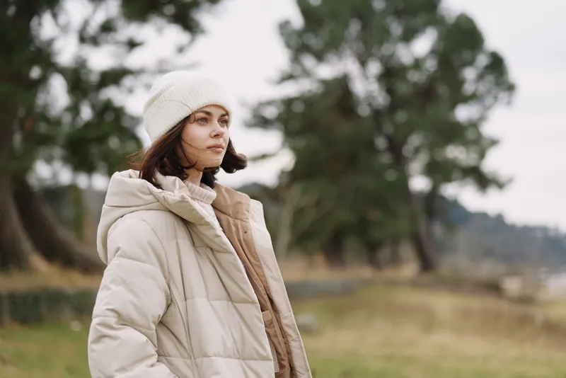 Woman in a beige puffer jacket and knit hat stands in a park setting, calm and thoughtful, capturing a peaceful outdoor moment with soft natural light and a blurred green background.