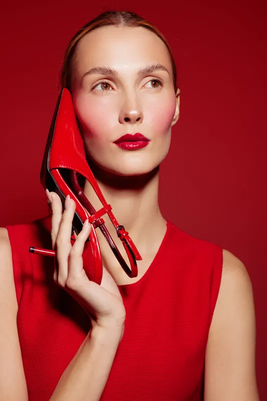 Stylish woman in red dress holds red high heel shoe near face against red background. Concept of fashion, beauty, elegance, and bold color contrast in studio portrait.