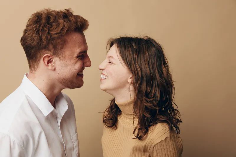 Smiling young couple facing each other closely, sharing a joyful moment. Casual clothing and warm beige background create a cozy, intimate atmosphere of happiness and connection.