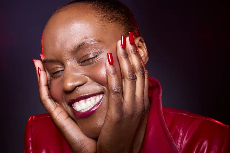 Close-up of a happy dark skinned African woman with short hair smiling with eyes closed, hands on face. She wears red nail polish and a red leather jacket on dark background.