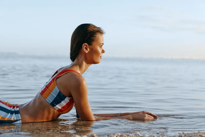 Woman beach ocean swimsuit exercise fitness wellness summer outdoors, side profile, woman kneeling in shallow water stretching, natural light, soft waves, peaceful horizon