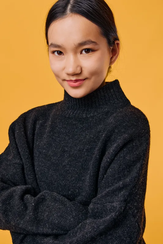 Young woman with dark hair wearing black sweater stands with crossed arms and gentle smile on orange background, looking confidently and warmly at camera in studio portrait.