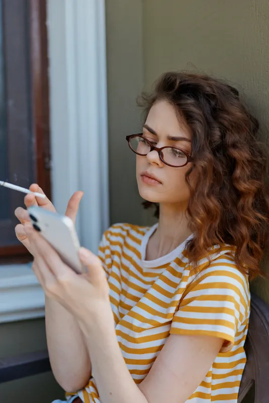 A thoughtful woman in a striped t shirt sits indoors, looking at her smartphone with glasses, creating a calm home lifestyle moment for social media and stock photography