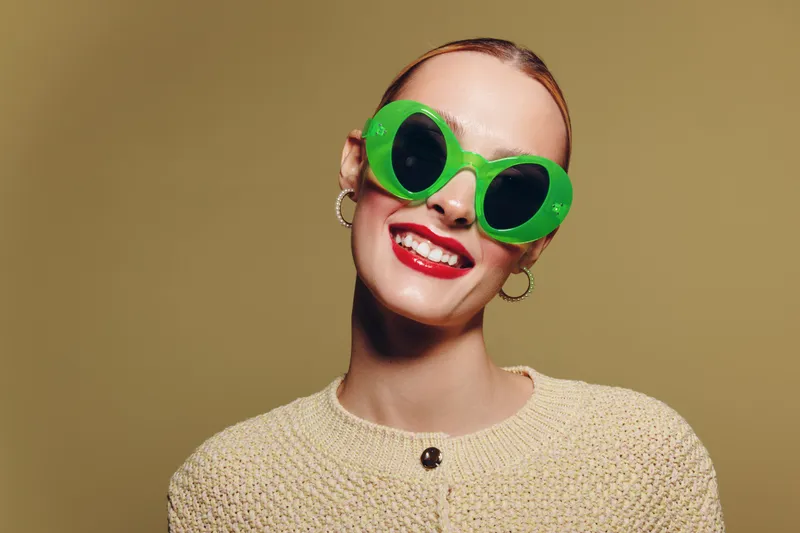 Smiling woman with bright red lipstick wears large green sunglasses and beige knitted sweater. Studio portrait on brown background. Concept of fashion, style, and joyful expression.