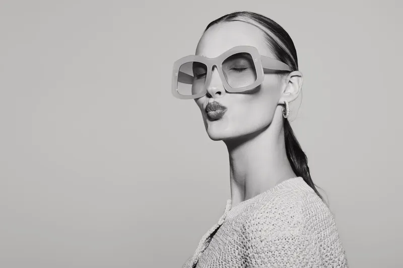 Young caucasian woman with fair skin wearing oversized sunglasses and textured sweater sending a kiss. Studio portrait in black and white with modern fashion style concept.