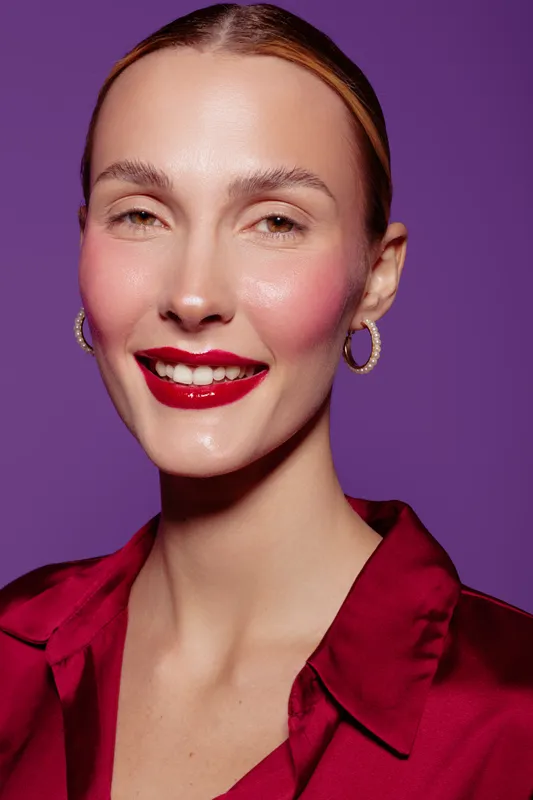 Smiling woman with red lipstick and hoop earrings poses against purple background. She wears a red satin blouse and has smooth skin and pulled-back hair. Beauty and fashion concept.