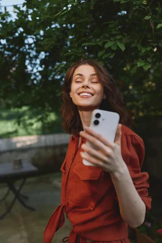 Smiling woman in a rust dress taking a selfie in a sunlit outdoor setting, with leafy trees behind. Casual lifestyle portrait conveying joy, confidence, and wellness