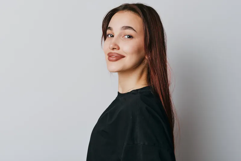 Young woman with long hair and natural makeup posing in black shirt against plain background. Portrait with confident smile and casual style for lifestyle or beauty concept.