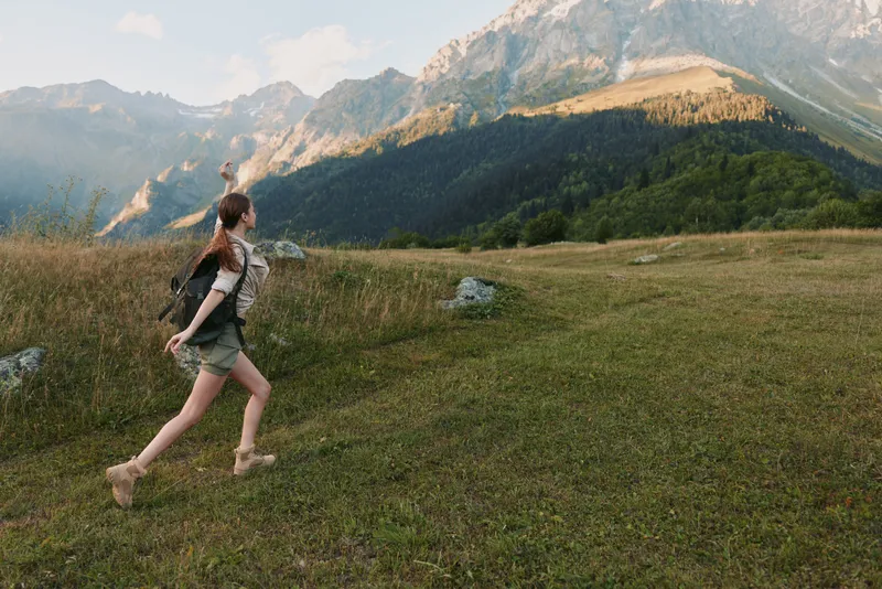 Young woman hiking in a grassy mountain meadow, exploring nature under blue skies, carrying a light backpack, and enjoying fresh air. Her casual outdoor outfit and sturdy boots convey a relaxed,