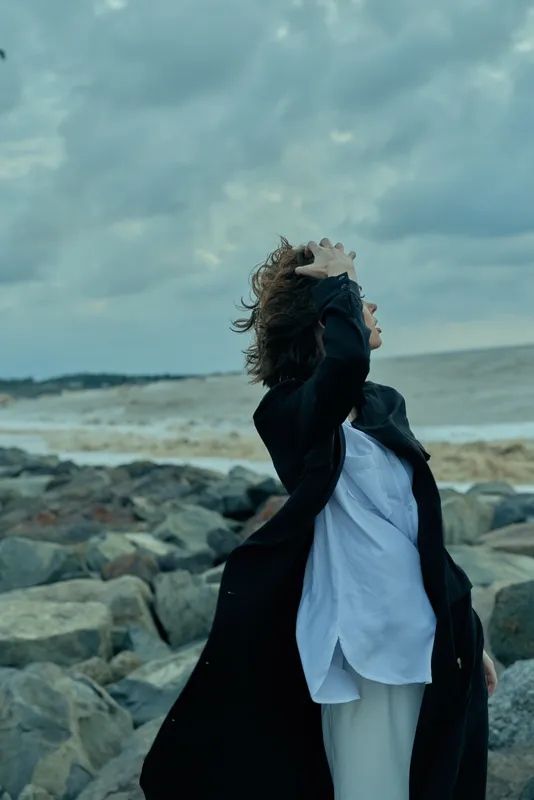 Dramatic wind swept scene with a woman on a rocky beach, black coat swirling, white shirt billowing, dramatic clouds over the sea, capturing movement and freedom