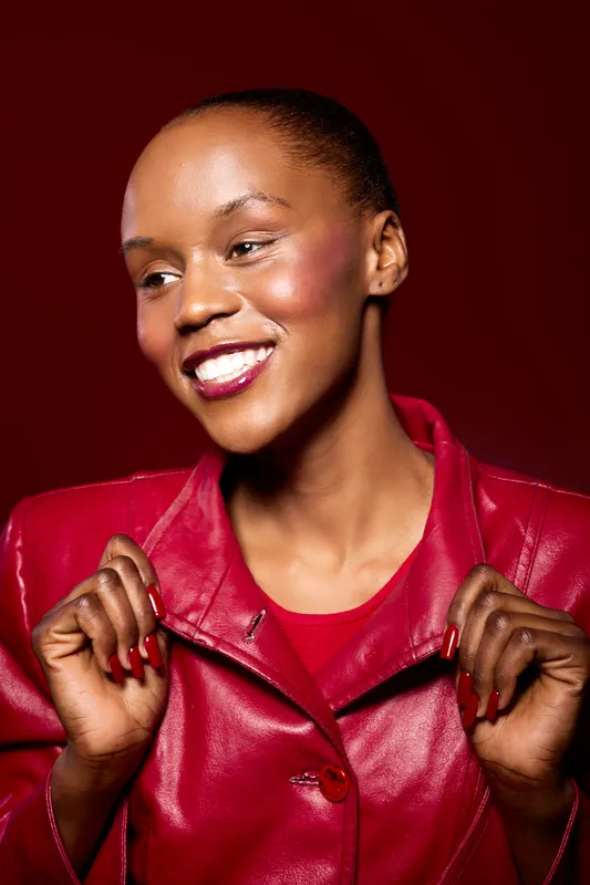 Smiling African woman with dark skin wearing red leather jacket and matching red shirt. She holds the collar of her jacket and looks away with a joyful expression on a red background.