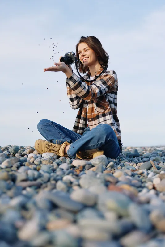 Woman sits on a pebble beach in casual plaid shirt and jeans, tossing stones into the air with joyful motion, capturing carefree outdoor play and bright light on a windy coast.