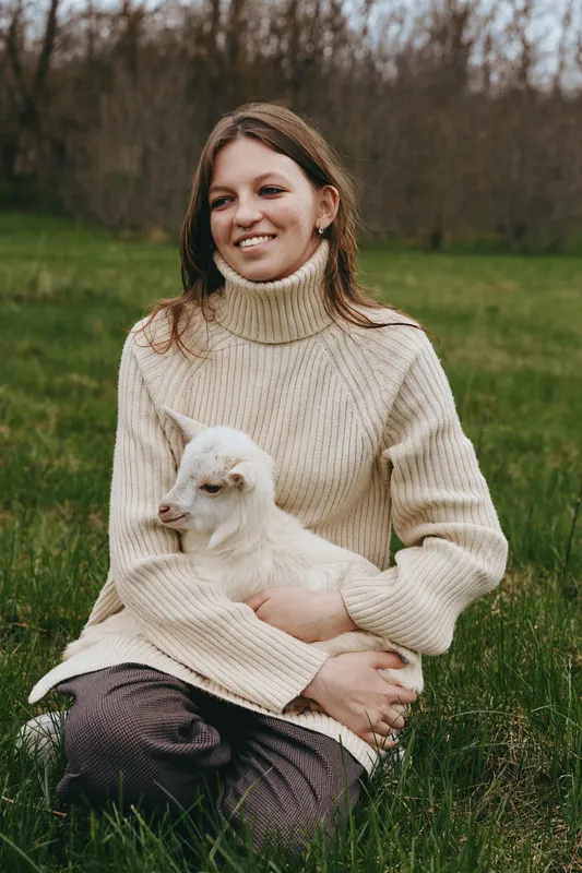 A young woman in a beige turtleneck sweater sits on grass holding a white baby goat while smiling gently in a natural outdoor setting during autumn.