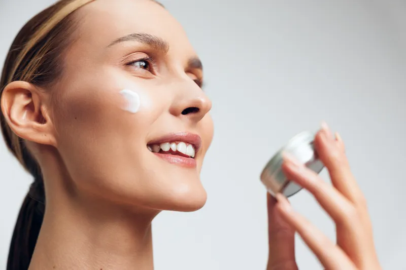 A smiling young woman applies moisturizer cream on her cheek holding a jar in hand. The closeup shows healthy skin and skincare routine concept with natural beauty and self care.