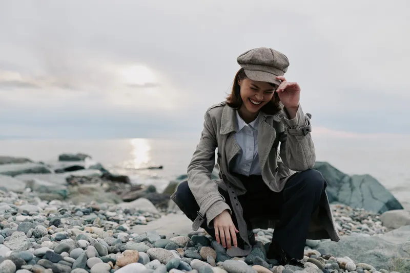 Smiling woman in vintage outfit crouches on rocky beach near water during cloudy day. Natural light highlights casual fashion and relaxed outdoor mood by the sea.