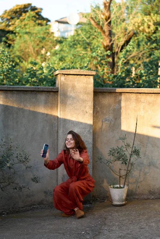 Cheerful woman outdoors, taking a selfie in a sunlit courtyard, wearing a red dress, relaxed pose, warm tones, soft breeze, lush greenery in the background, candid joy