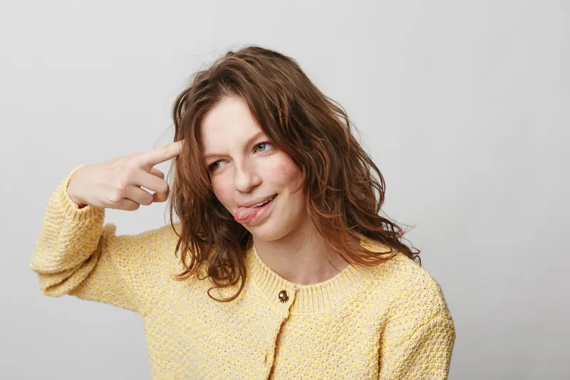 Young woman with curly hair in yellow sweater makes funny face and points finger to head. Isolated on white background. Concept of humor, playfulness, and silly expression.
