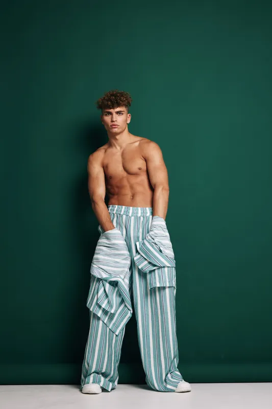 Man, shirtless, with toned muscular body wearing loose striped pants and holding matching shirt with boxes in studio. Male model with curly hair posing casual in fashionable clothes on green