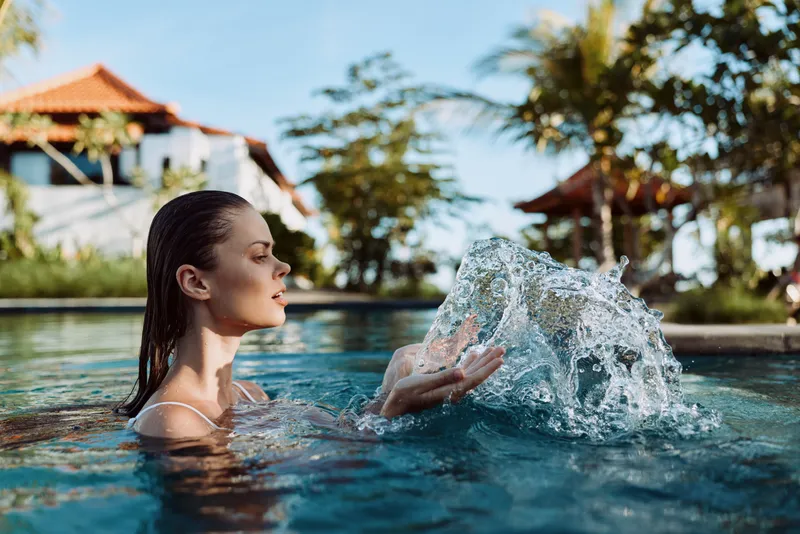 Active woman in a pool splashes water, bright outdoor moment with sunlit glow, relaxed pose, clear water shimmer, warm breeze, natural colors, serene lifestyle